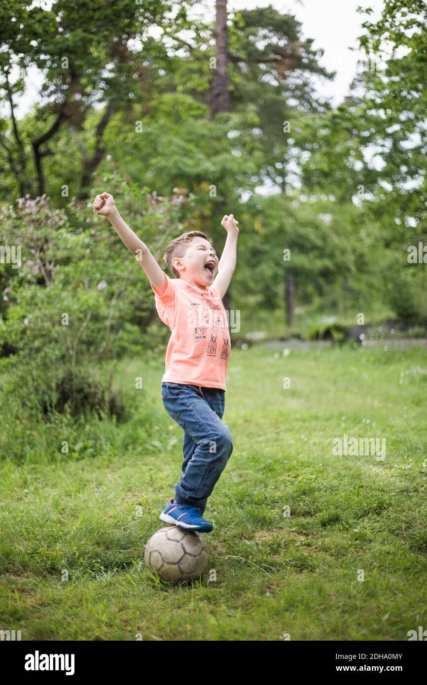 Aufgeregter Junge, der mit erhobenen Armen auf dem Fußball steht Ball im Hinterhof Stockfoto