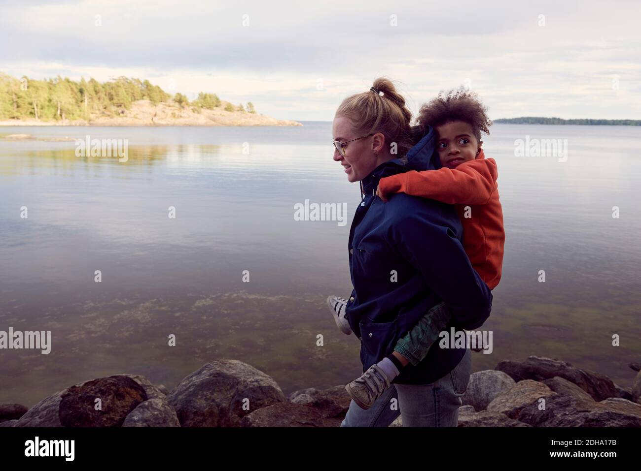 Seitenansicht der jungen Mutter, die dem Sohn Huckepack gibt Seeufer Stockfoto