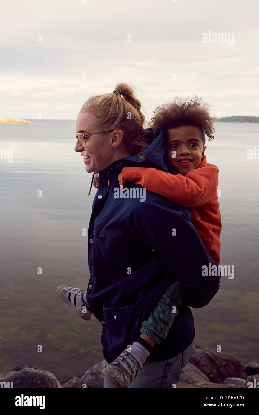Seitenansicht der lächelnden Mutter, die dem Sohn Huckepack gibt Seeufer Stockfoto