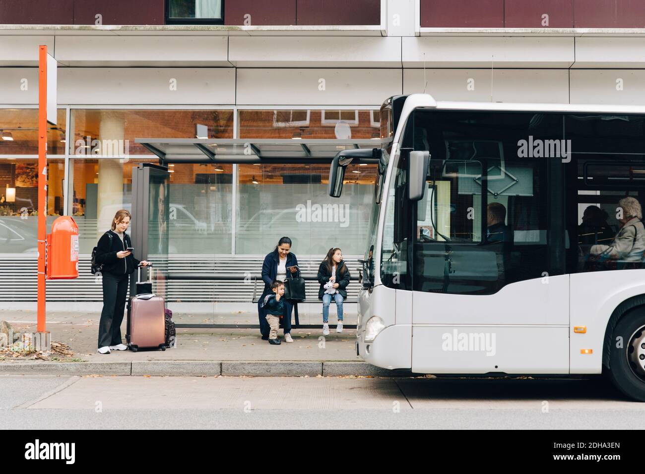 Volle Länge der Frau mit Gepäck und Familie wartet auf Bus an der Bushaltestelle in der Stadt Stockfoto