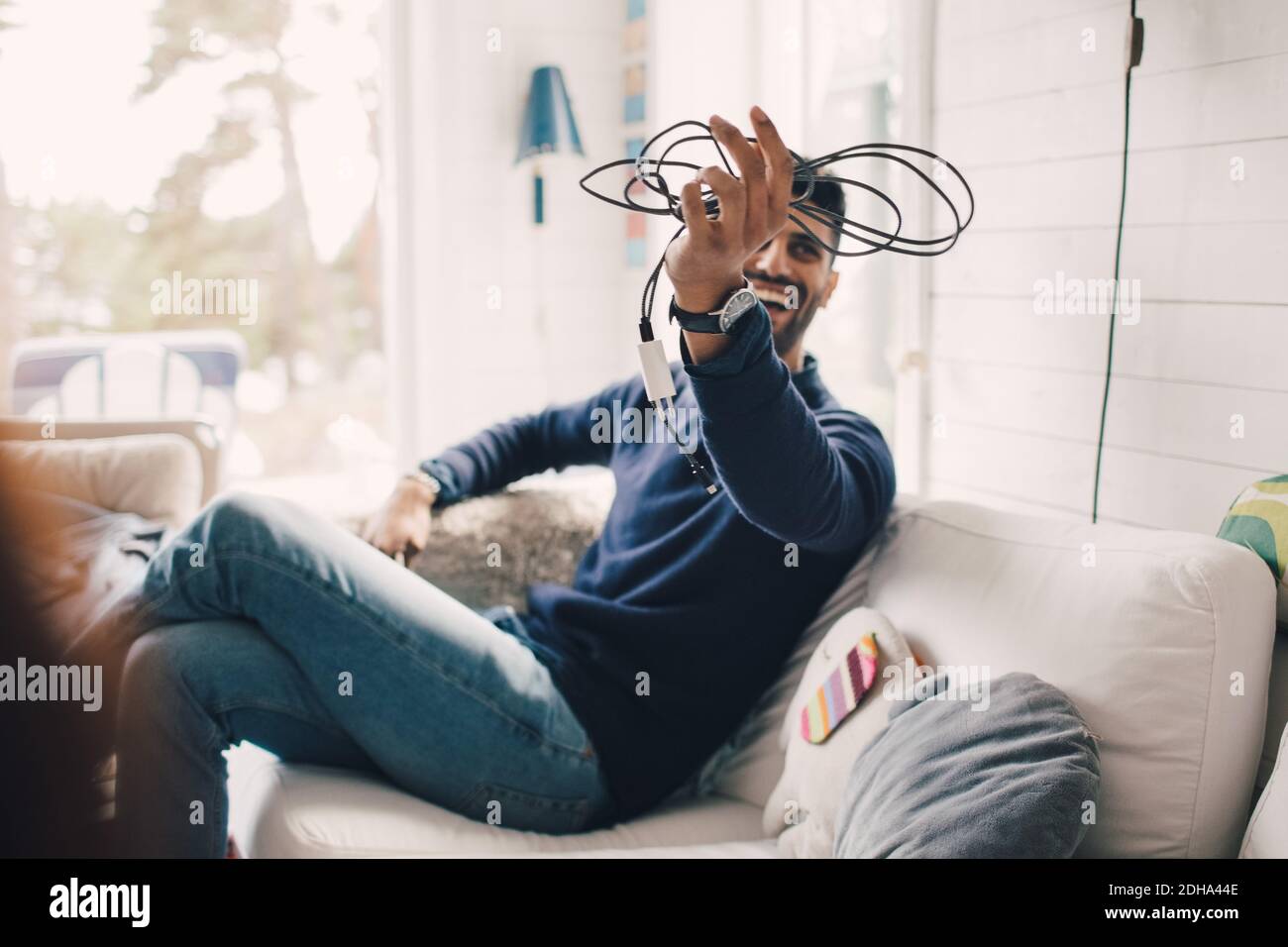 Ein fröhlicher Mann, der ein Elektrokabel in der Hand hielt, während er auf dem Sofa saß Zu Hause Stockfoto