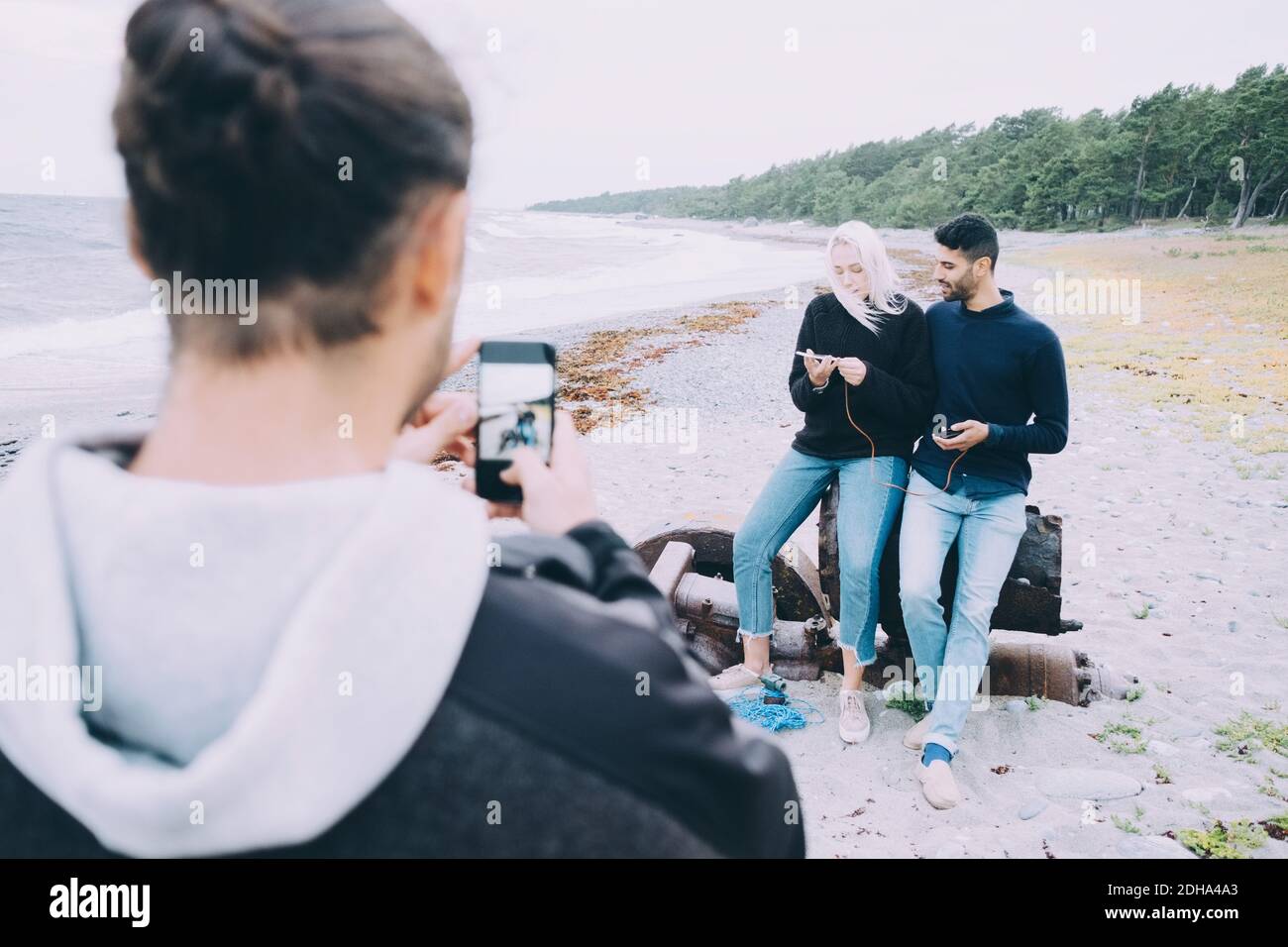 Rückansicht eines jungen Mannes, der Freunde fotografiert, die auf Metall sitzen Am Strand Stockfoto