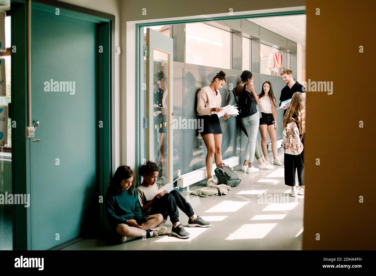 Mädchen und Junge sitzen in der Tür, während Professor im Gespräch mit Schüler im Schulkorridor Stockfoto