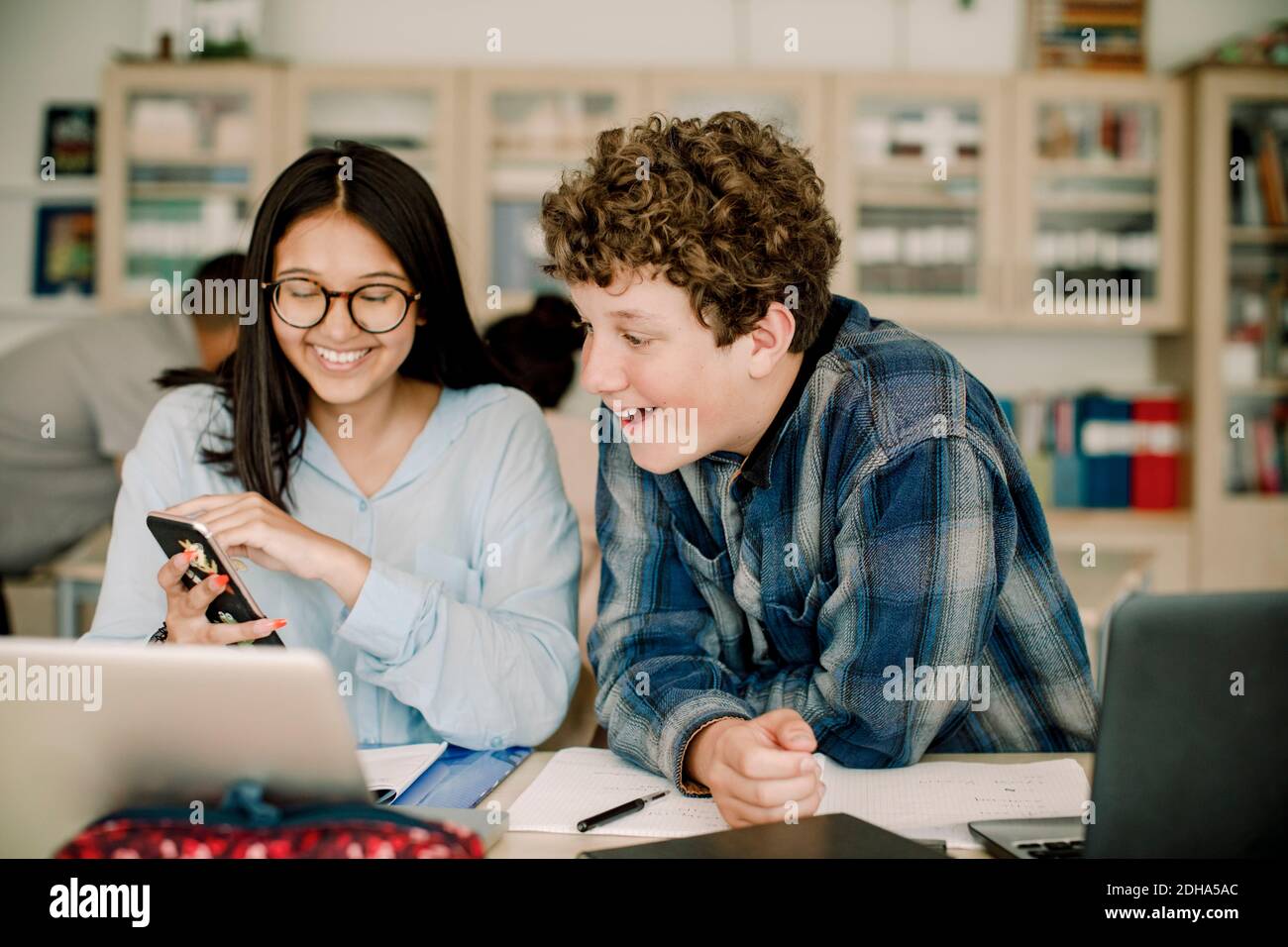 Lächelndes Teenager-Mädchen zeigt Smartphone zu männlichen Freund, während Sitzen im Klassenzimmer Stockfoto