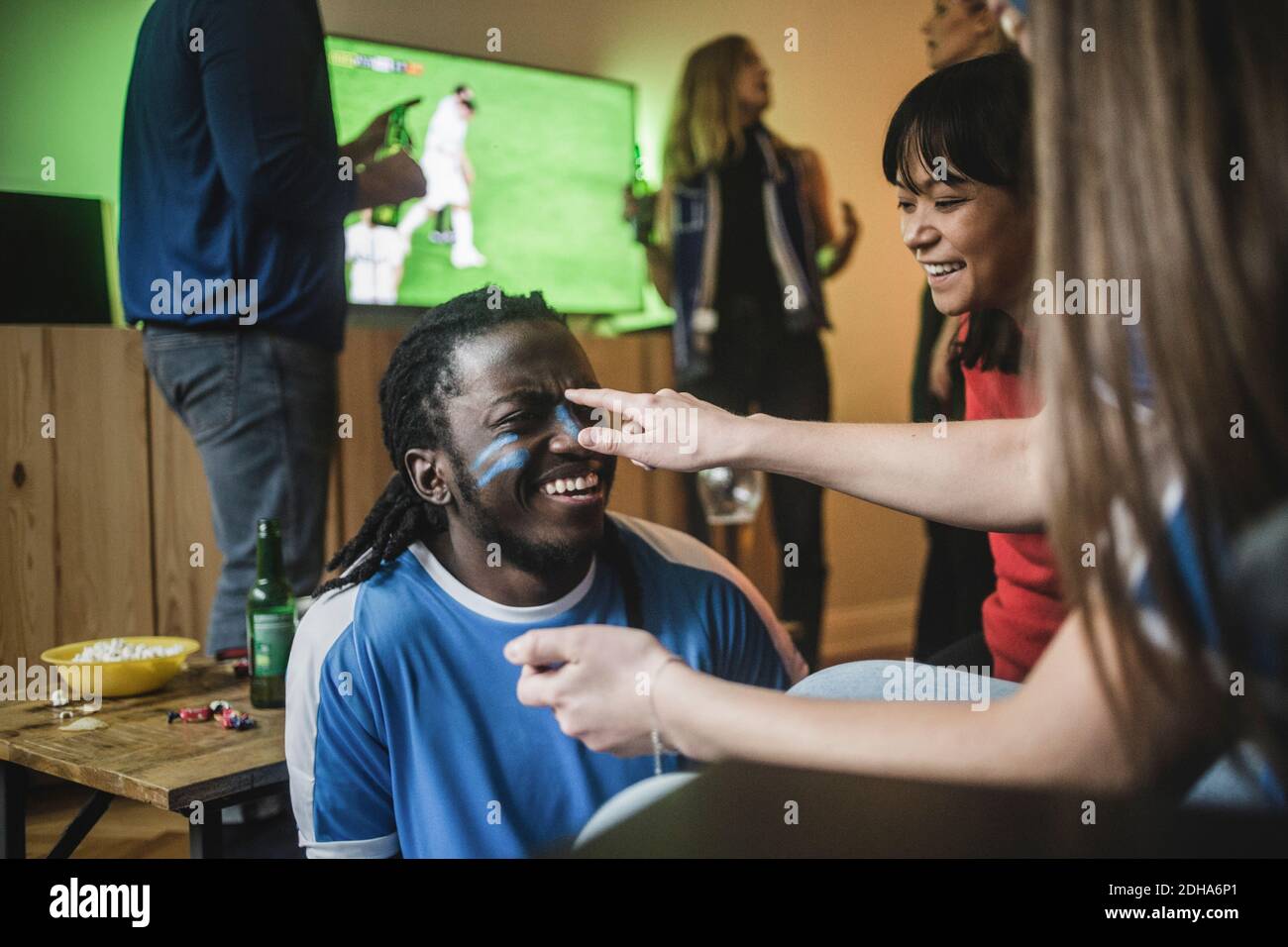 Junge Frau, die während des Fußballs Gesichtsfarbe auf die Wange eines Freundes auftragen Übereinstimmung Stockfoto