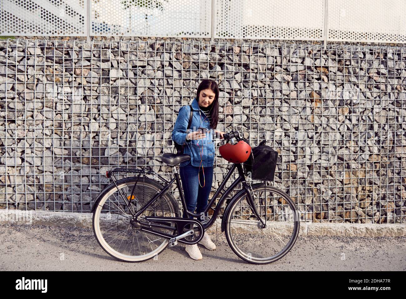 Volle Länge der Frau mit Smartphone während des Stehens mit Fahrrad gegen Steinmauer Stockfoto