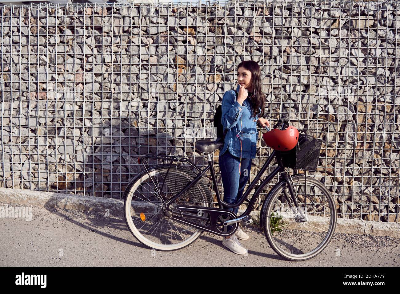 Volle Länge der Frau, die weg schaut, während sie mit Fahrrad steht Gegen Steinmauer Stockfoto