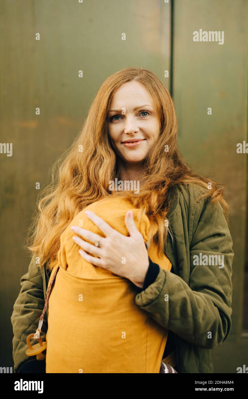 Porträt einer lächelnden Mutter, die das Baby hält, während sie gegen die Wand steht Stockfoto