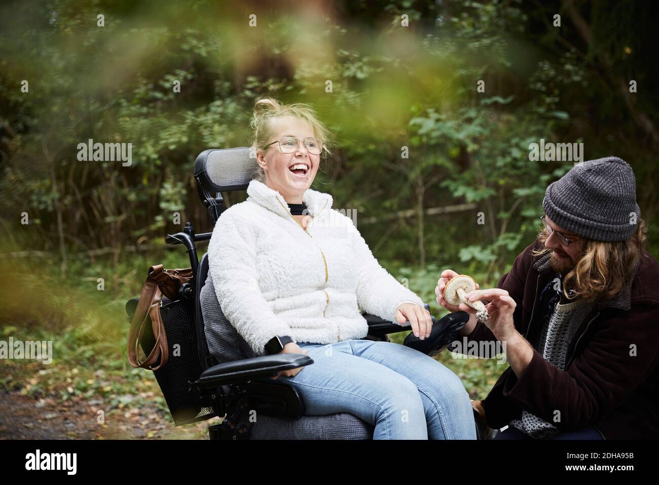 Fröhliche behinderte Frau im Rollstuhl von Hausmeister hält Pilz an Wald Stockfoto