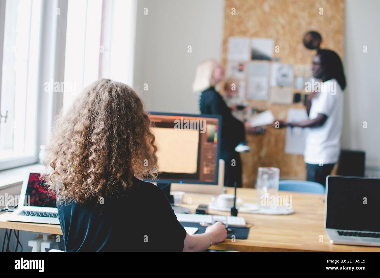 Rückansicht der jungen Geschäftsfrau mit Computer am Tisch mit Kollegen im Hintergrund im Creative Office Stockfoto