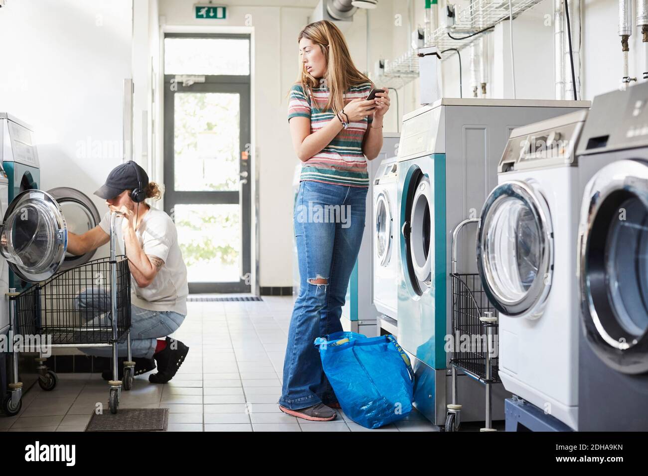 Frau mit Handy, während der Mann Wäsche im Waschsalon macht Stockfoto