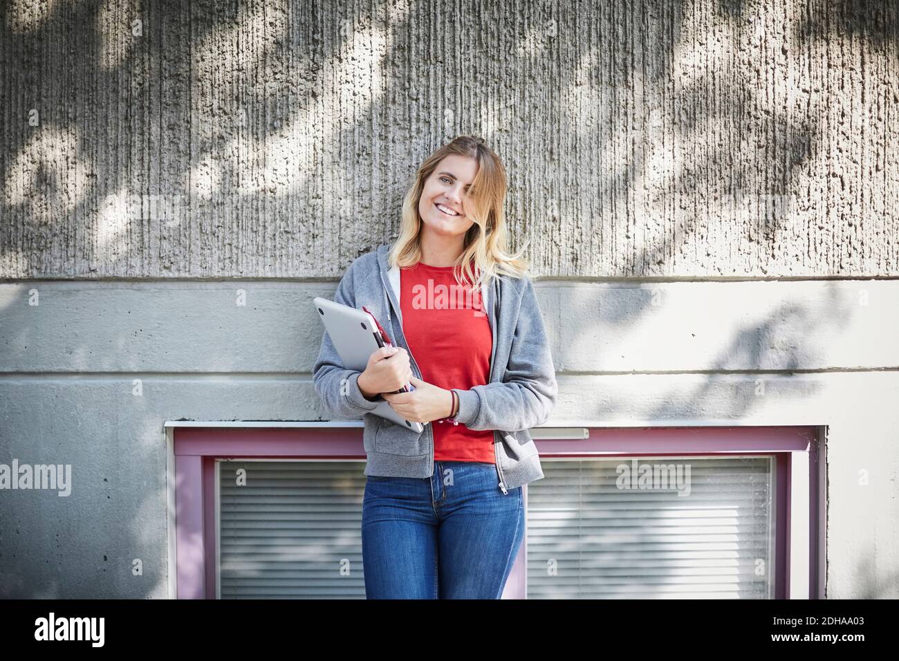 Porträt eines lächelnden Studenten, der einen Laptop in der Hand hält, während er sich dagegen hält Wand an sonnigen Tag Stockfoto