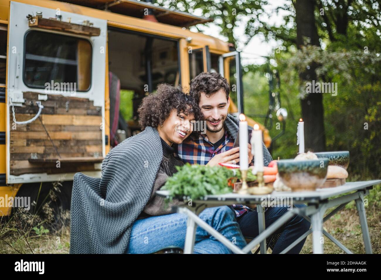 Lächelndes junges Paar, das sich das Handy teilt, während es in eine Decke gehüllt ist Zusammen beim Camping im Wald Stockfoto