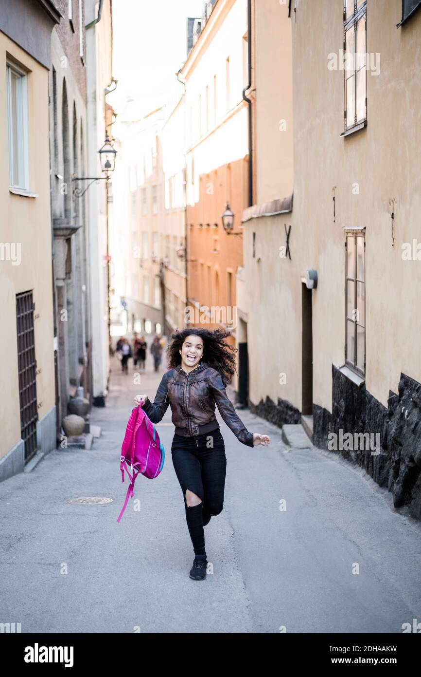 Volle Länge der glücklich Teenager-Mädchen läuft mit Rucksack auf Straße inmitten von Gebäuden Stockfoto