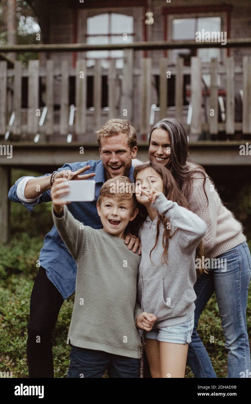 Lächelnder Junge nimmt Selfie mit der Familie auf Handy, während Steht gegen Haus Stockfoto