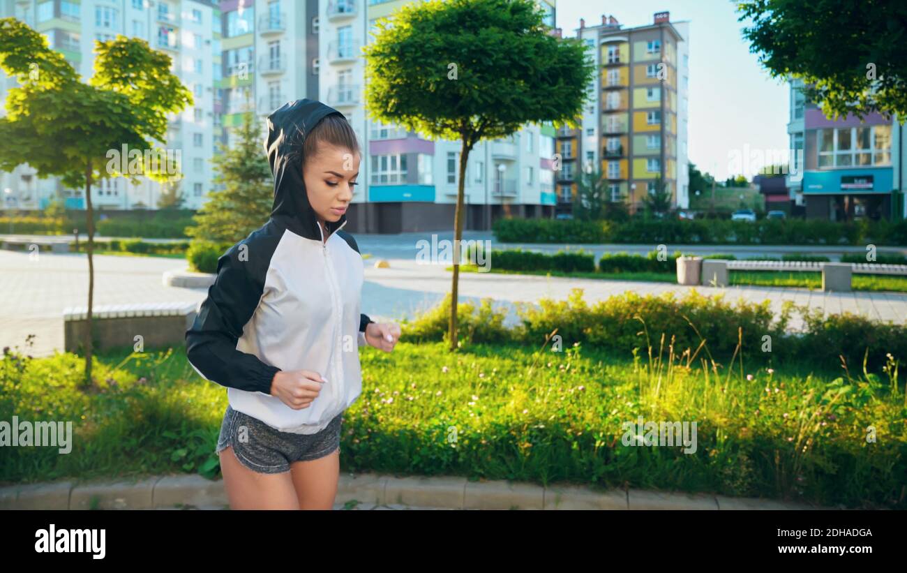 Junge Frau trägt Shorts und Kapuze auf dem Kopf Joggen im sonnigen Sommermorgen in den Straßen. Atemberaubende weibliche Läuferin macht Cardio-Training im Freien, Multisto Stockfoto