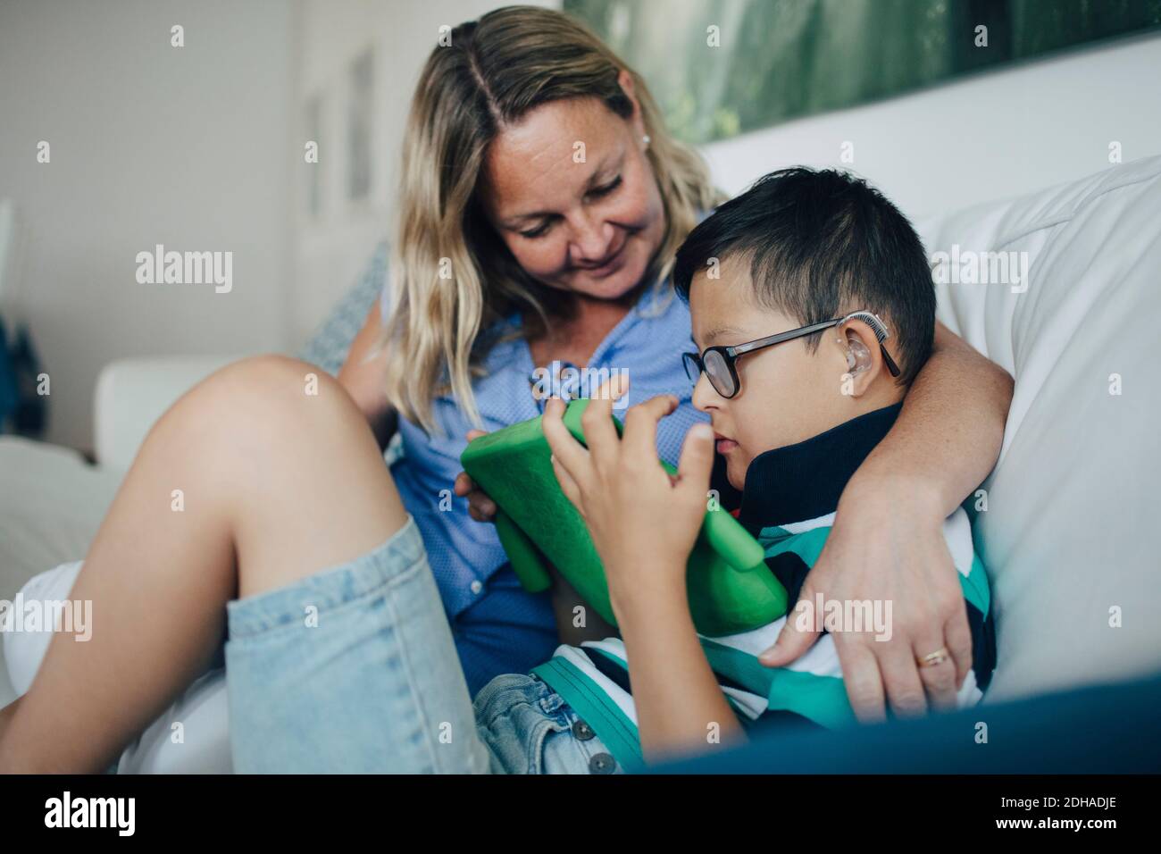 Lächelnde Mutter schaut auf behinderten Sohn mit digitalen Tablet, während Auf dem Sofa sitzen Stockfoto