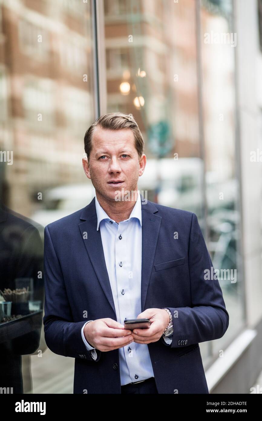 Erwägen reifen Geschäftsmann mit Smartphone wegschauen, während stehend Durch Glasfenster Stockfoto