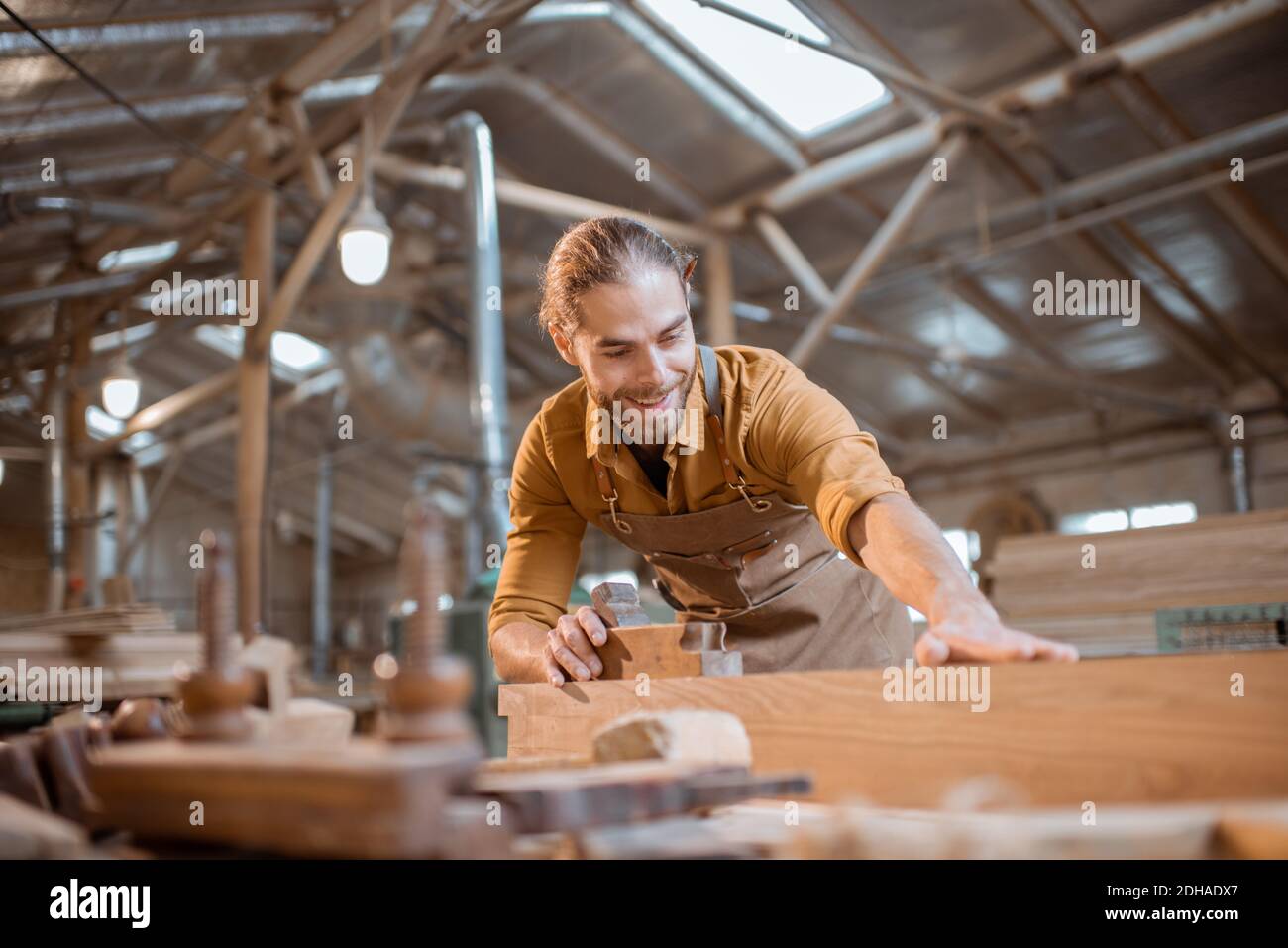 Hübscher Zimmermann, der mit einem Holz arbeitet, auf die Bar schaut, die Qualität in der Werkstatt überprüft Stockfoto