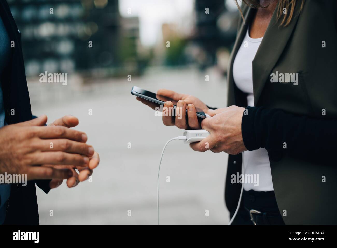 Mittelteil der Geschäftsfrau, die im Stehen mit einem Kollegen das Mobiltelefon auflädt In der Stadt Stockfoto