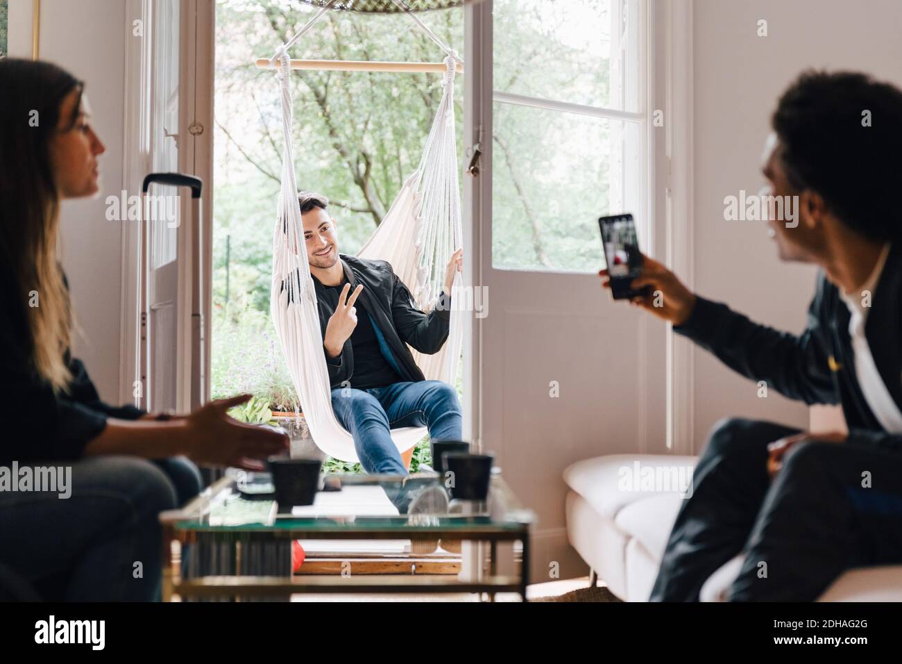 Mann fotografiert Freund mit Smartphone, während er im Schaukel sitzt Zu Hause Stockfoto