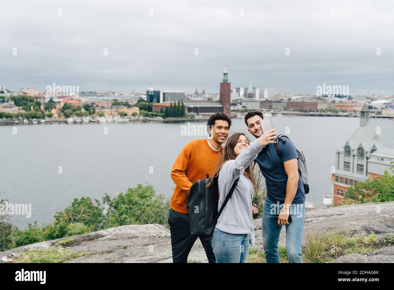 Lächelnde Freunde, die Selfie mit dem Mobiltelefon machen, während sie sich im Stehen befinden Fluss in der Stadt gegen Himmel Stockfoto