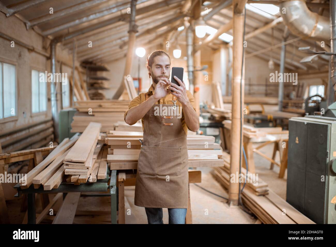 Schöner Zimmermann im Gespräch auf Smartphone oder mit dem Telefon, um einige Holzarbeiten in der Schreinerei zu entwerfen. Konzept der mobilen Technologien in der Zimmerei Stockfoto
