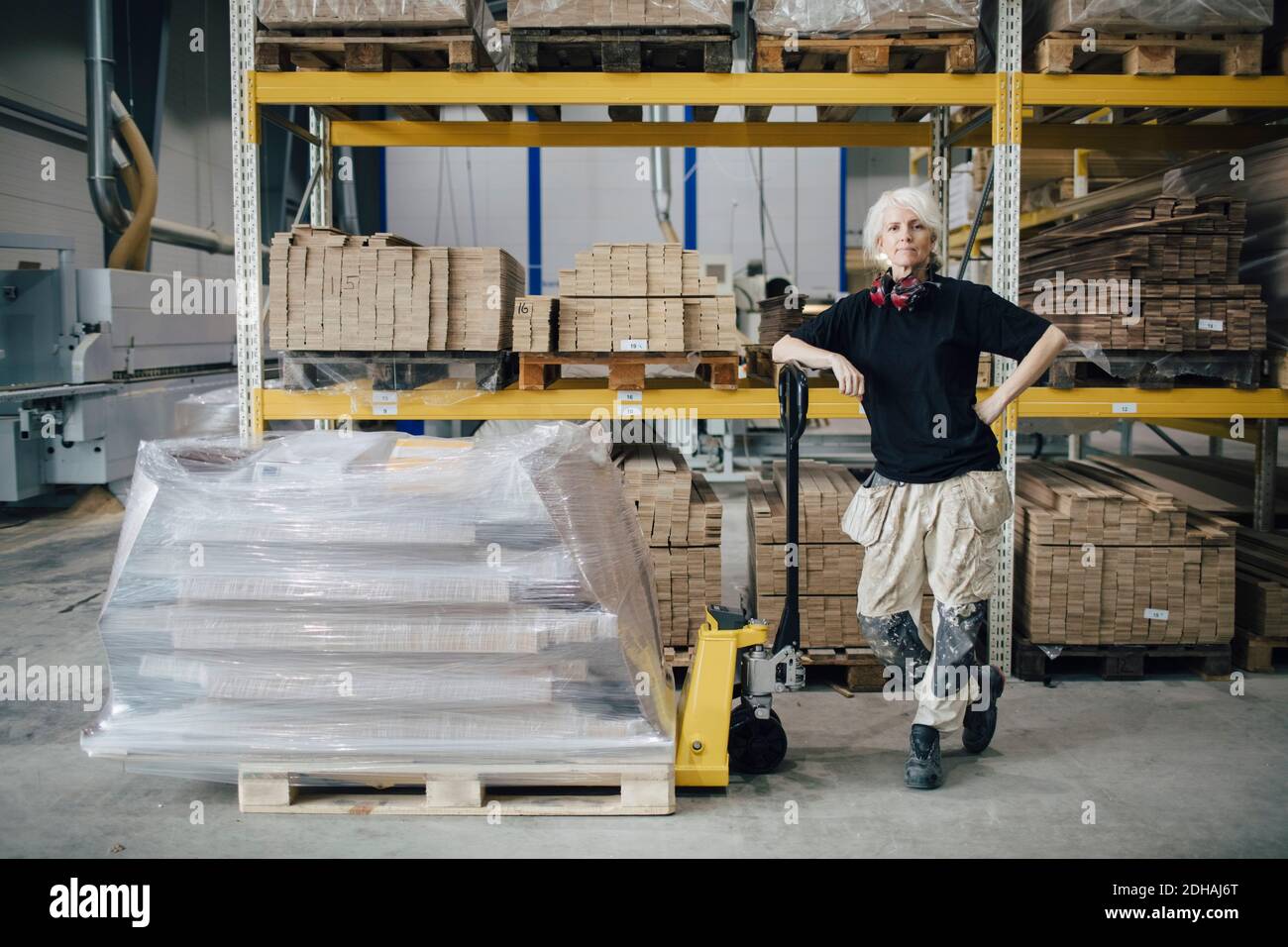 Volle Länge des selbstbewussten Arbeiters, der an gestapelten Brettern steht Palettenhubwagen in der Industrie Stockfoto