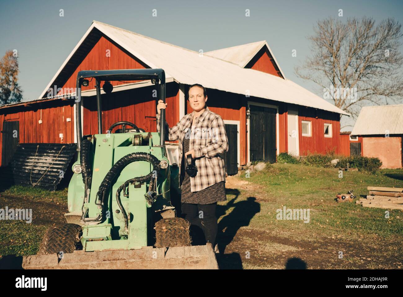 Porträt einer mittleren erwachsenen Farmerin, die neben einem landwirtschaftlichen Fahrzeug steht Gegen die Scheune Stockfoto