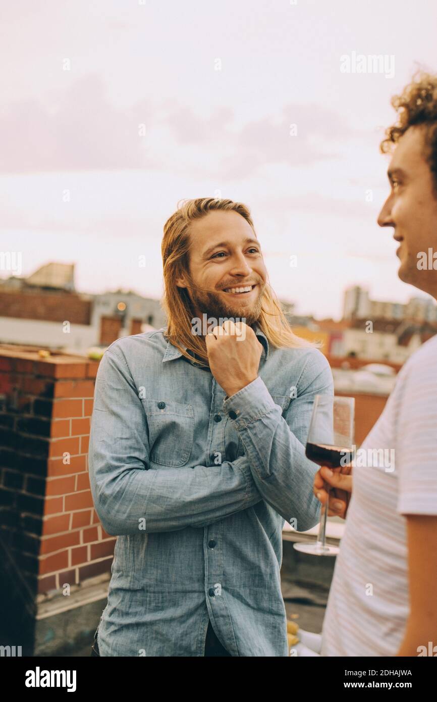 Lächelnde männliche Freunde reden, während sie auf der Dachterrasse stehen Party Stockfoto