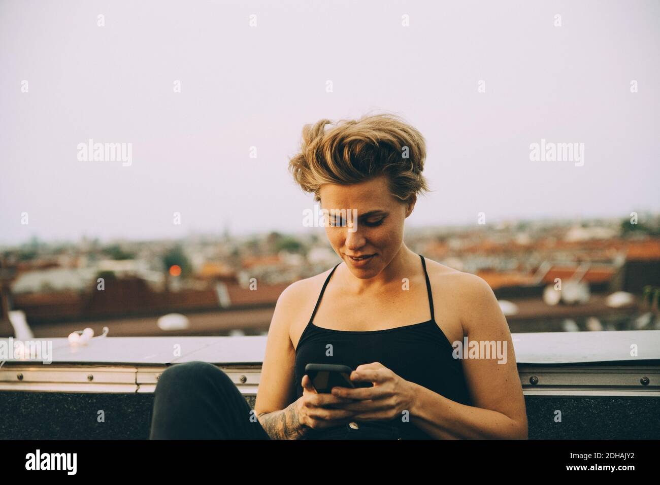 Lächelnde Frau mit kurzen Haaren Textnachrichten auf dem Smartphone Während Sie auf der Terrasse gegen den Himmel sitzen Stockfoto