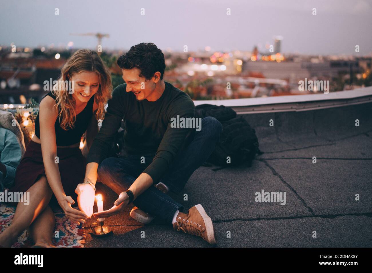 Lächelndes junges Paar schützt brennende Kerze mit Händen während des Sitzens Auf der Terrasse gegen den Himmel in der Dämmerung Stockfoto