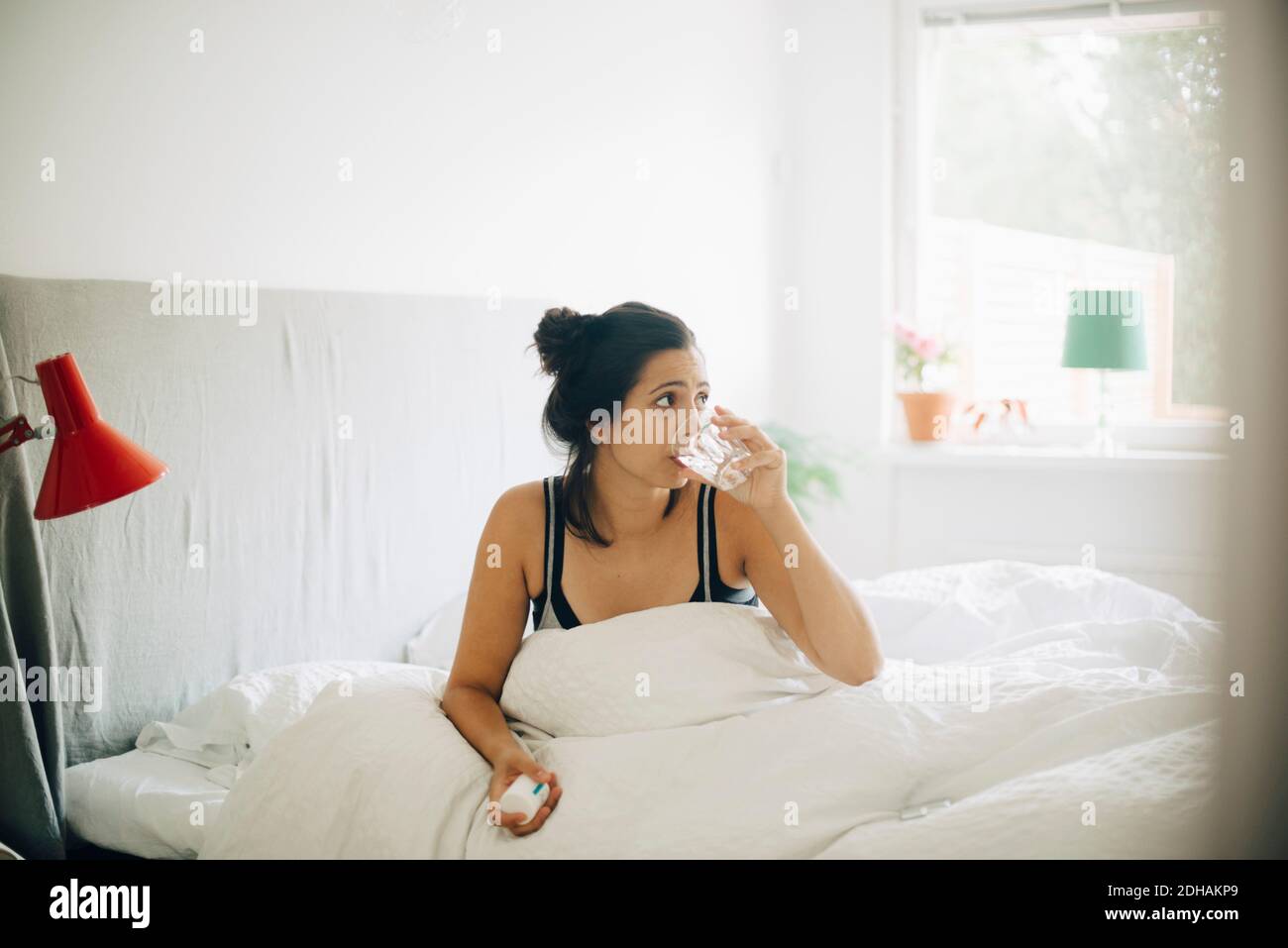 Frau, die Wasser trinkt, während sie zu Hause Pillen auf dem Bett hält Stockfoto