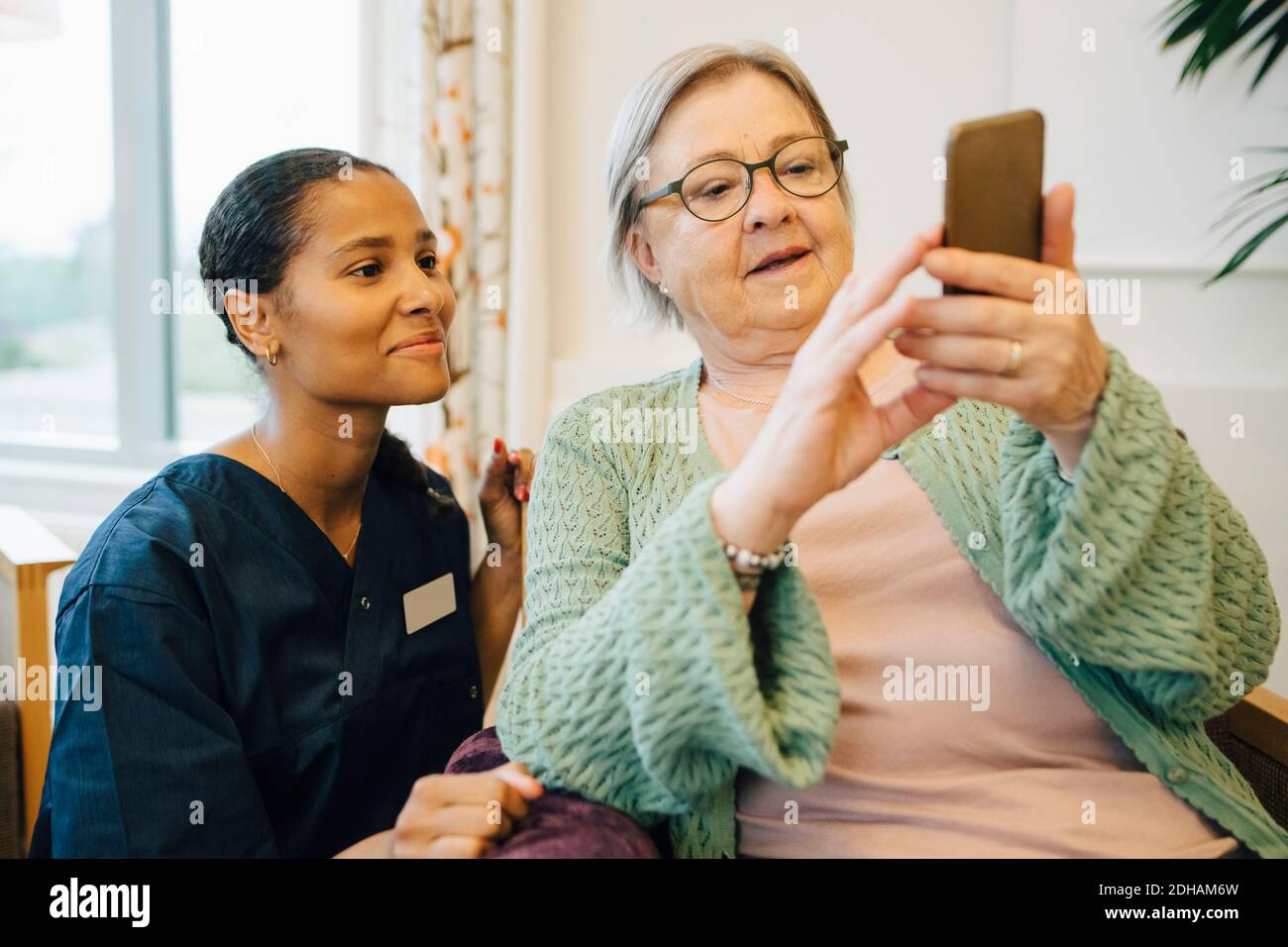 Lächelnde Hausmeisterin, die ältere Frau mit dem Handy ansieht Im Pflegeheim Stockfoto