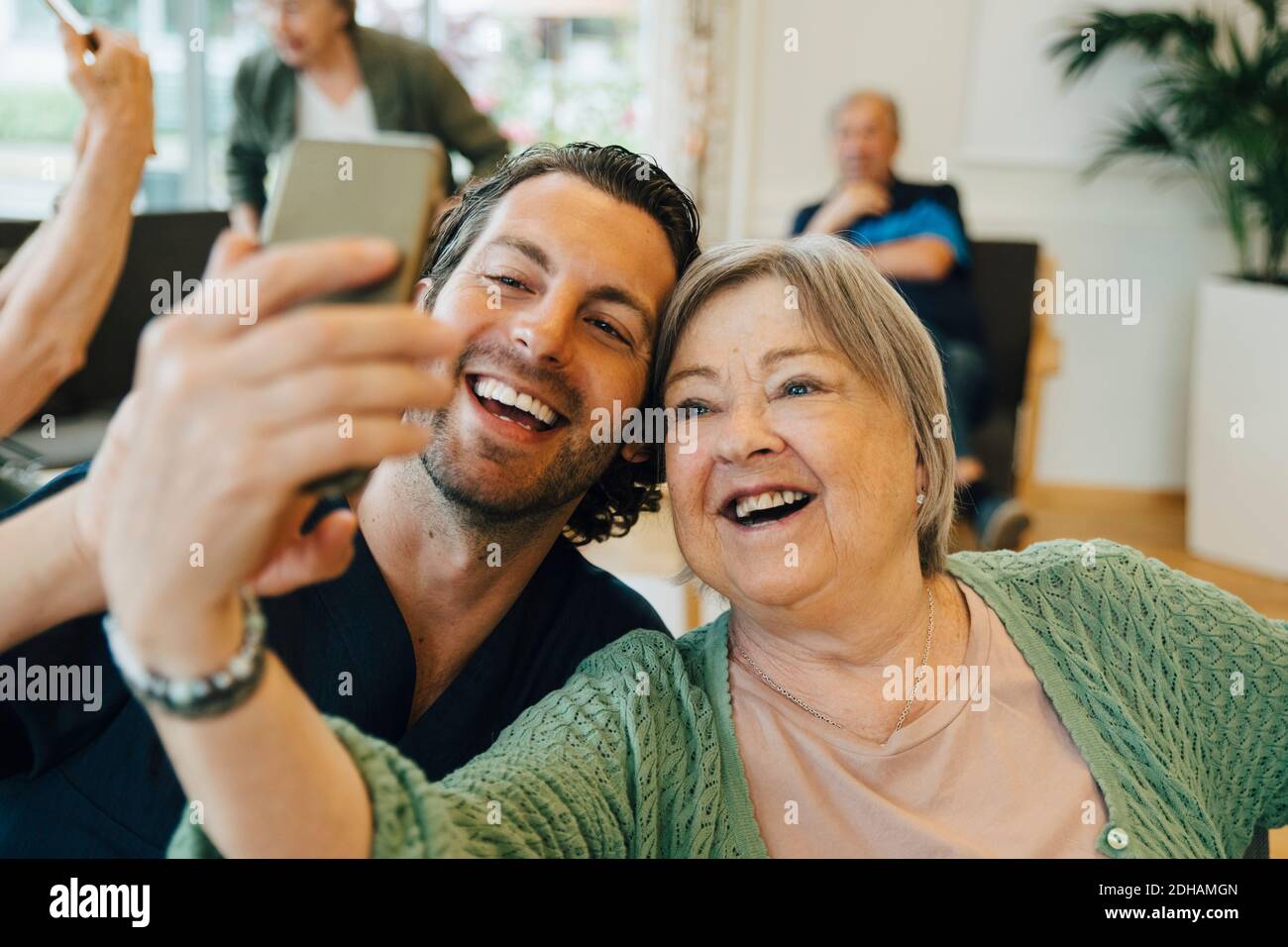 Lächelnde ältere Frau, die Selfie mit männlichen Gesundheitshelfer nimmt, während Sitzen im Altersheim Stockfoto