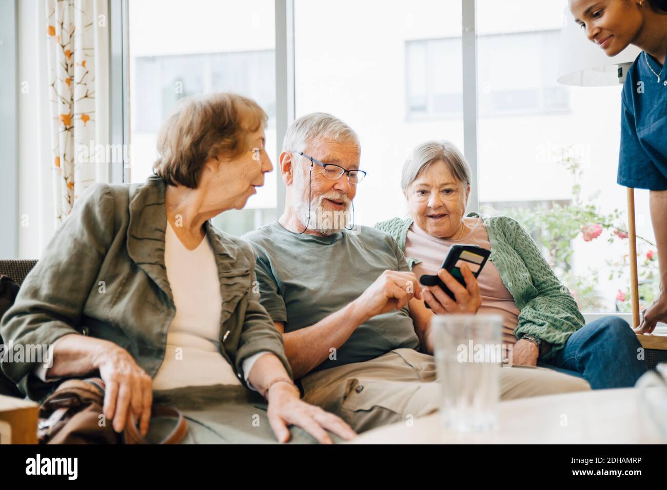 Älterer Mann, der Smartphone benutzt, während er inmitten von Frauen sitzt Altersheim Stockfoto