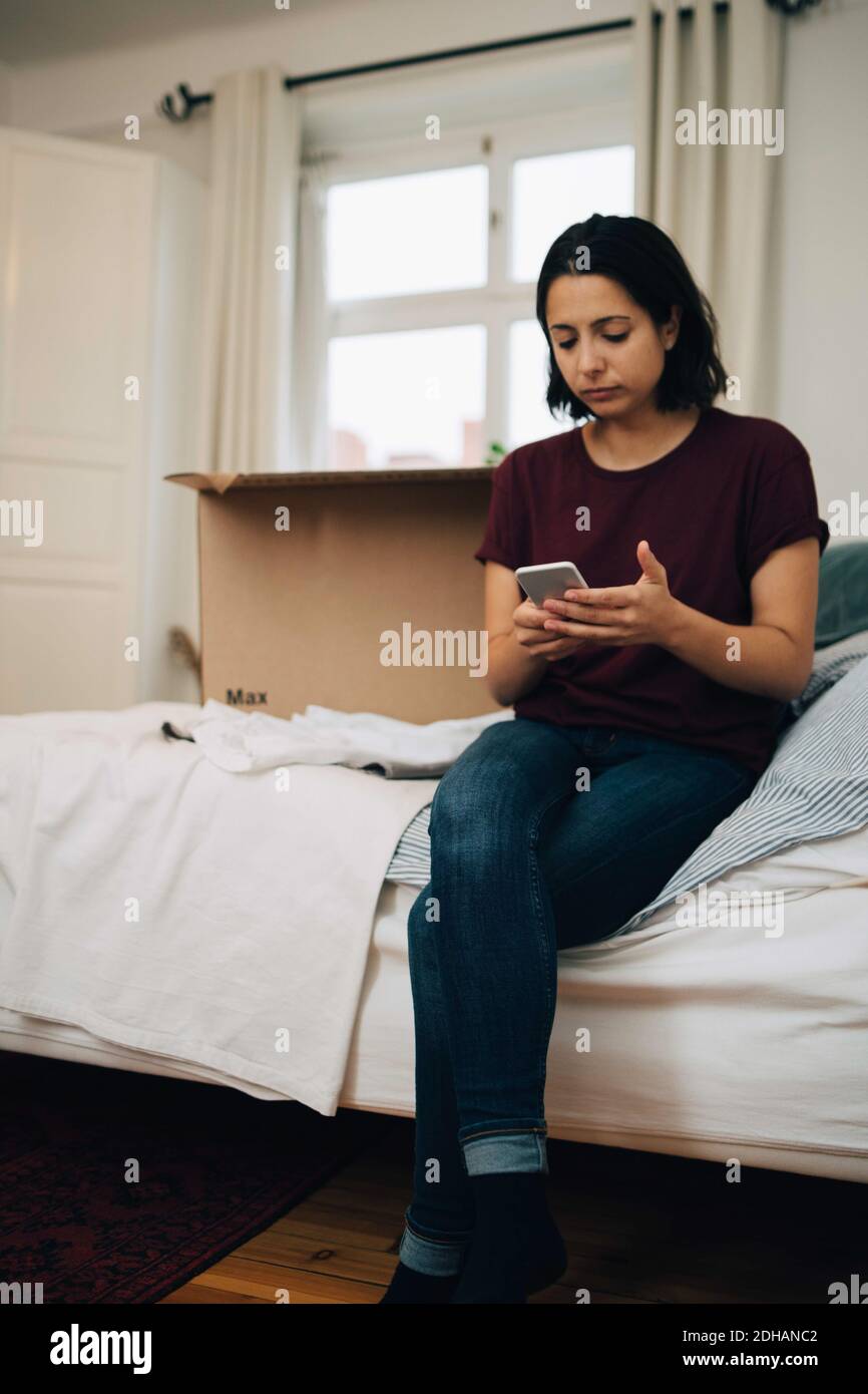 Frau mit Handy, während sie an Box auf dem Bett sitzt Stockfoto