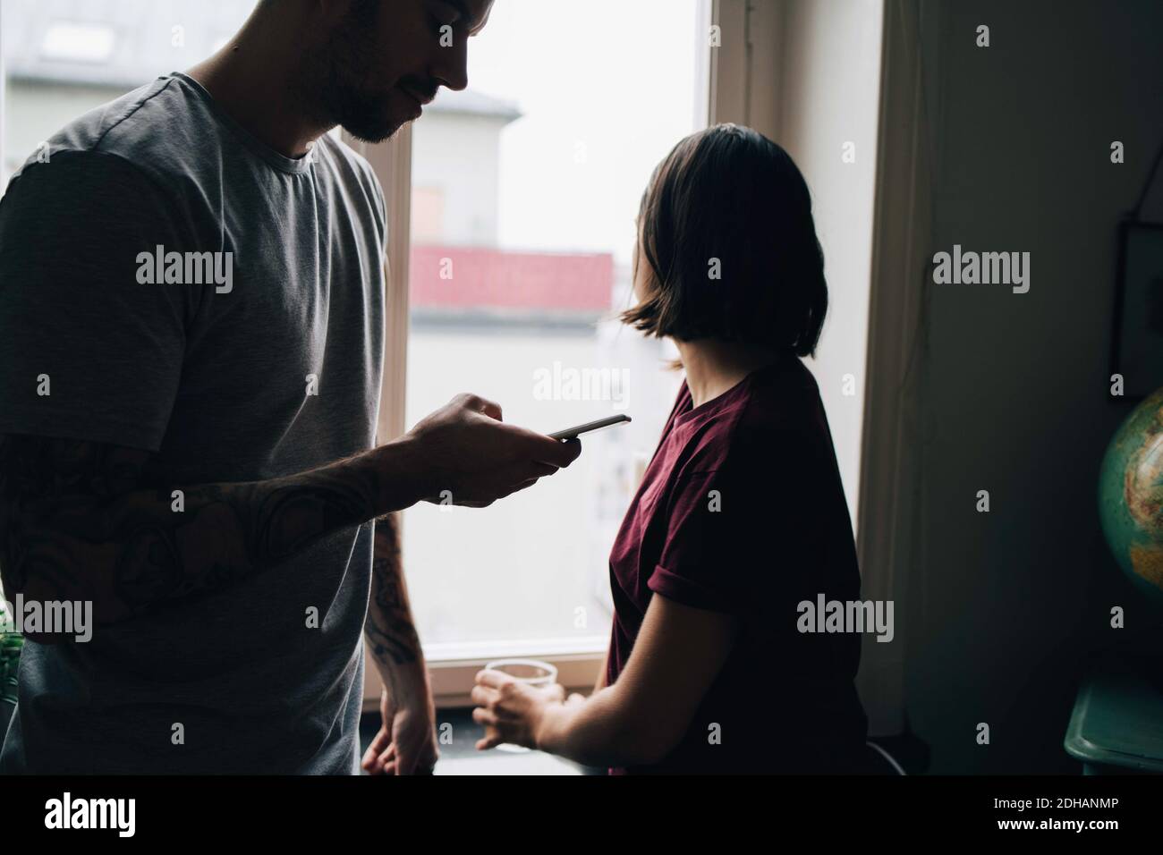 Frau schaut durch das Fenster, während Mann Handy benutzt Zu Hause Stockfoto