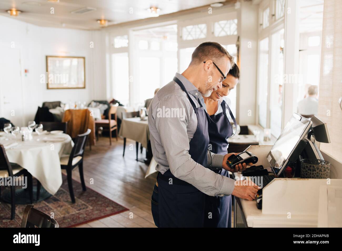 Lächelnde Besitzer, die den Computer benutzen, während sie den Kreditkartenleser in der Hand halten Restaurant Stockfoto