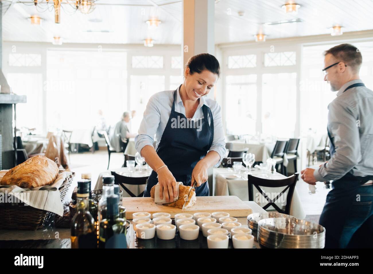 Lächelnder Besitzer schneidet Brot auf dem Tisch, während Mitarbeiter im Hintergrund Stockfoto