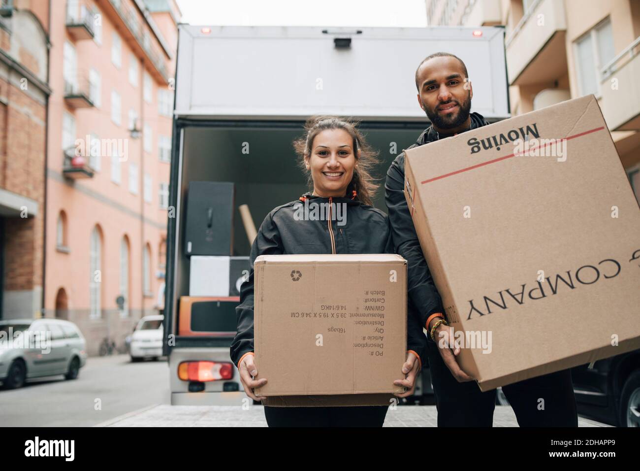 Portrait von männlichen und weiblichen Boten tragen Pappkartons während Stehen gegen Lieferwagen in der Stadt Stockfoto