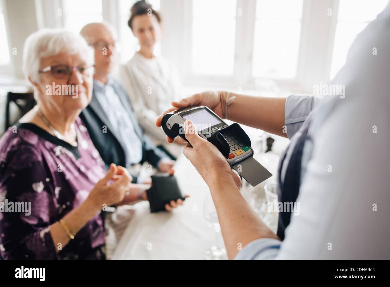 Mittelteil des Besitzers mit Kreditkartenleser, während älterer Mann Und Frauen sitzen am Tisch im Restaurant Stockfoto