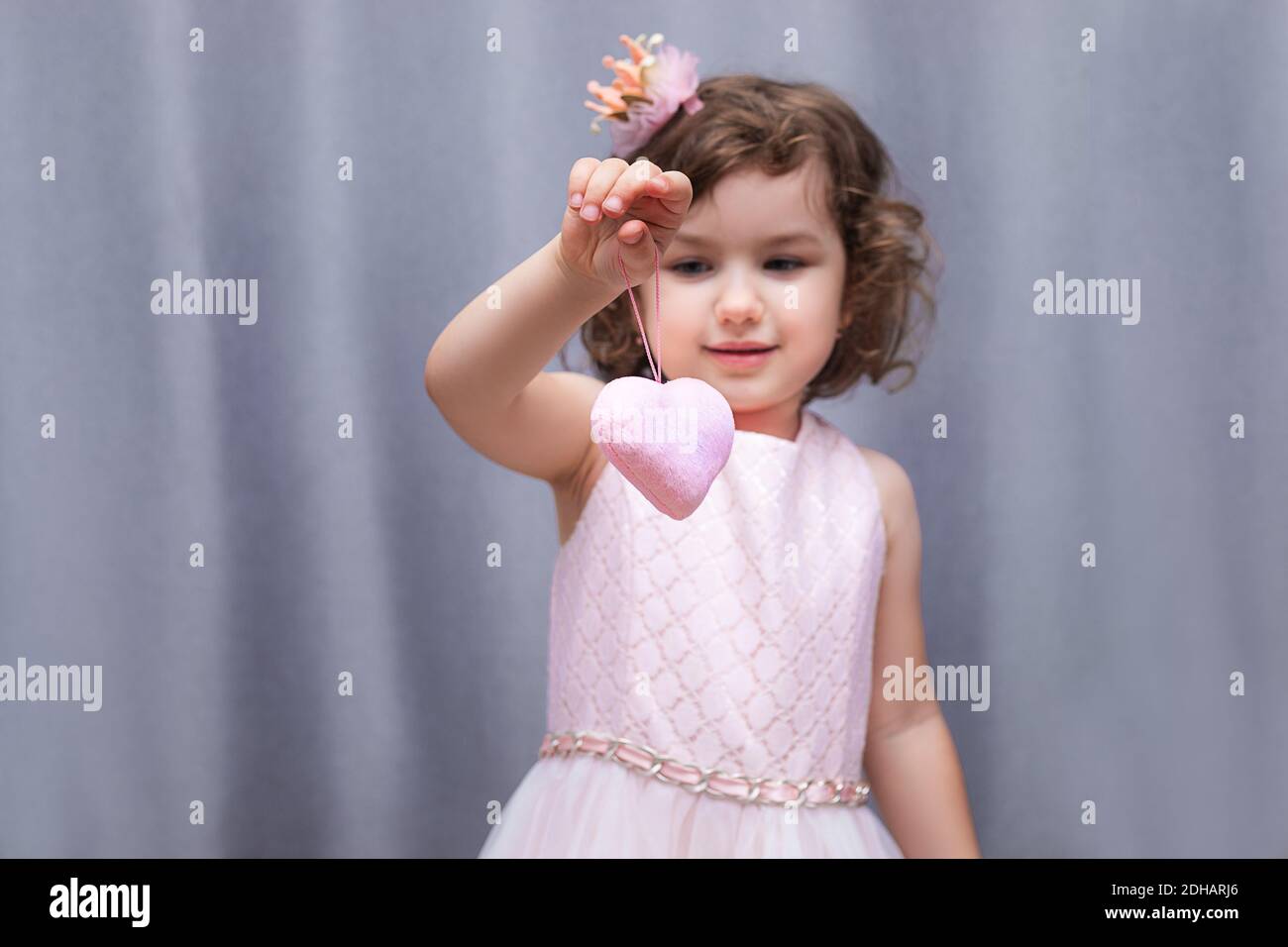 Ein rosa weiches Spielzeug in Form eines Herzens in den Händen eines 4-5-jährigen Mädchens. Valentinstag zum Valentinstag. Nahaufnahme im Hochformat auf grauem Hintergrund Stockfoto