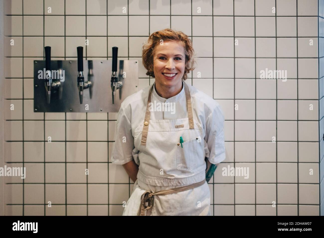 Porträt einer lächelnden Küchenchefin, die an der Wand mit weißen Kacheln steht In der Restaurantküche Stockfoto