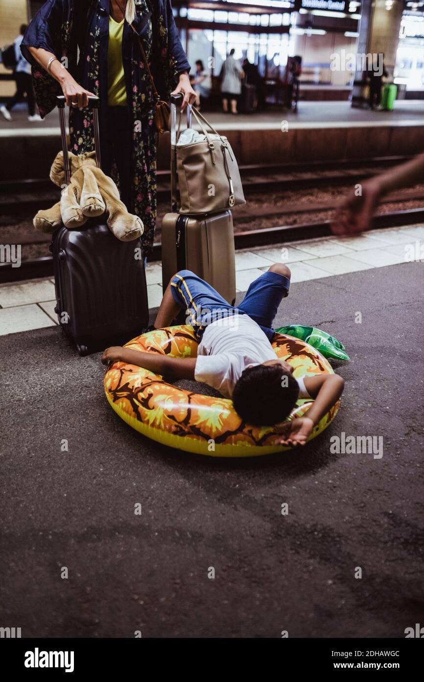 Junge liegt auf aufblasbarem Ring, während Mutter mit Gepäck steht Am Bahnhofssteig Stockfoto