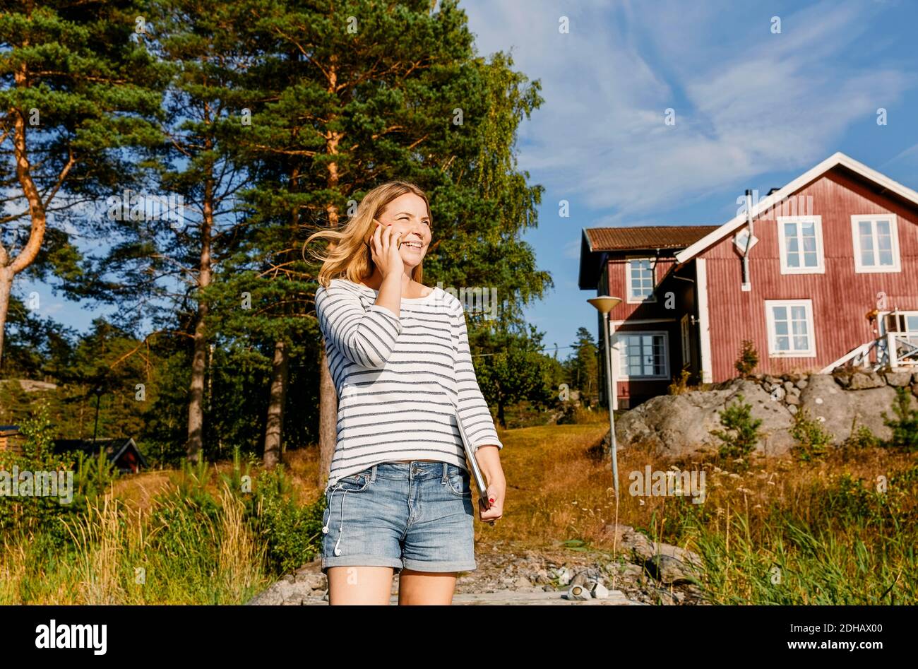 Lächelnde Frau mit mittlerem Erwachsenenalter, die im Stehen auf dem Mobiltelefon spricht Gegen Bäume und Haus Stockfoto
