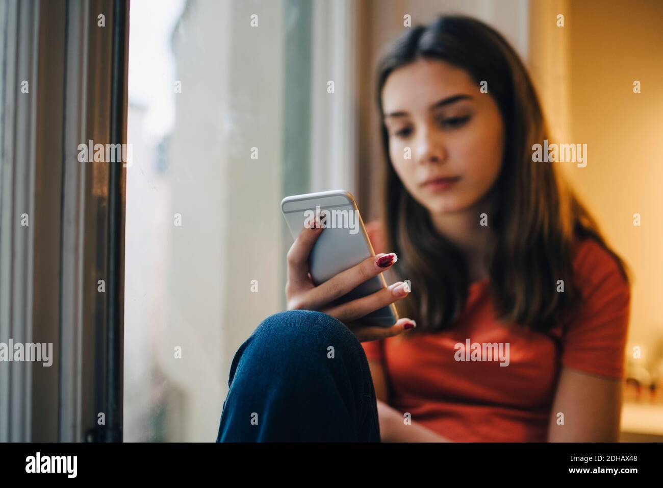 Nachdenklich Mädchen mit Smartphone, während auf Fensterbank sitzen Zu Hause Stockfoto