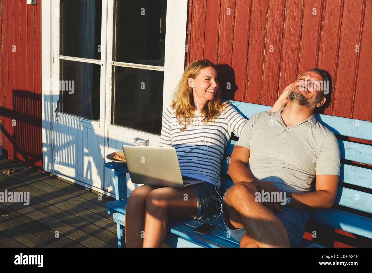 Glückliche Frau mit Laptop, während mit Mann auf Bank spielen Im Sommer auf der Veranda Stockfoto