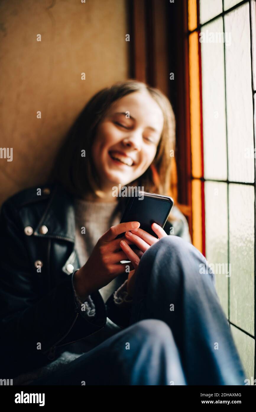 Lächelndes Mädchen, das Smartphone benutzt, während es am Fenster sitzt Zu Hause Stockfoto