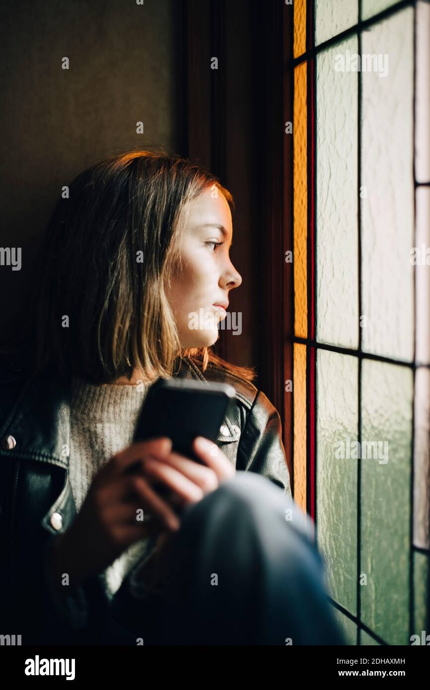 Nachdenkliches Mädchen mit Smartphone, das durch das Fenster schaut, während es sitzt Im Haus Stockfoto